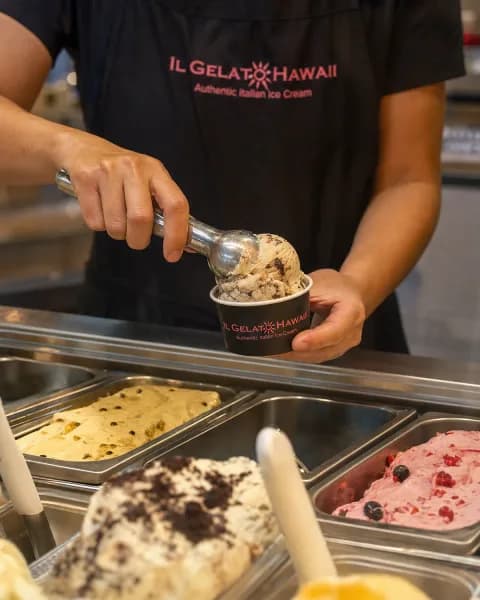 Apron-wearing employee mid-scoop, pulling a cookies-and-cream gelato from a steel display tub.