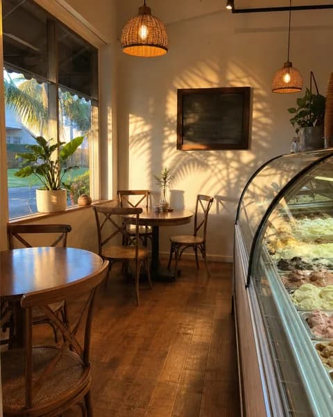 Empty cafe interior with a curved gelato display case, wooden tables, woven pendant lamps, and warm late-afternoon light pouring through the front window.