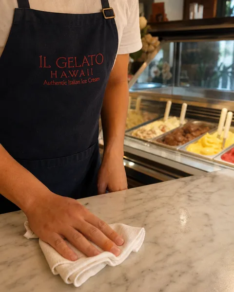 An employee in an IL GELATO HAWAII apron wiping down the marble counter, hands and brand mark in frame, no face.